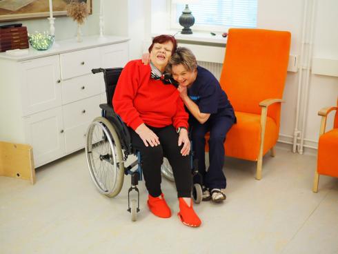 En äldre kvinna i en rullstol och en kvinnlig vårdare lutar mot varandra och skrattar gott.