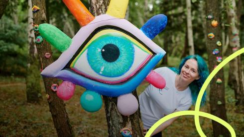 En leende blåhårig ungdom tittar fram bakom ett ögonformat miljökonstverk. 