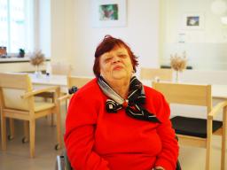 En äldre kvinna i en röd tröja sitter i vardagsrummet på en rehabiliterande bedömningsenhet.